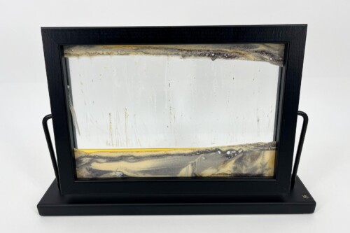 Black-framed glass art piece on a black pedestal, with amber-brown marbled bands and drips inside the panel with side handles visible on the frame.