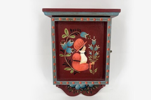 Decorative wooden cabinet door with a red panel showing a person in a red hat among floral motifs; teal frame with orange trim and dotted border.