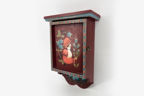 Decorative wooden wall cabinet with teal trim and orange dots, featuring a painted woman in a hat among plants on its front panel