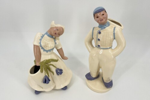 Pair of ceramic figurines: a seated woman in a cream dress with blue trim bending toward a flower vase, and a standing man in a cream outfit with a blue cap and blue accents.