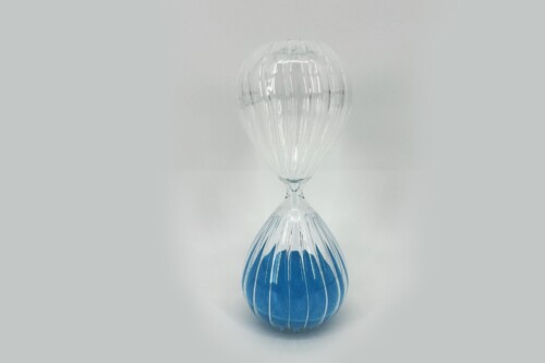 Clear glass hourglass with blue sand, standing upright on a white surface.