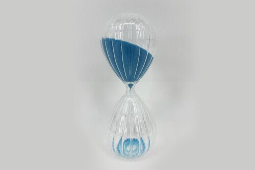 Hourglass with blue sand, blue-filled bottom chamber, on a reflective surface with a pale gray background.