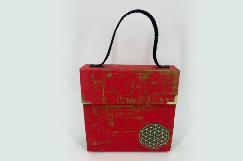 Red wooden decorative box with a black arched strap handle and brass corner accents, featuring a circular geometric emblem on the front.