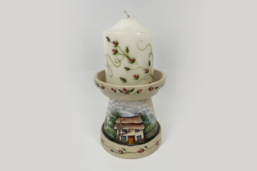 White decorative pillar candle with pink flower vines resting on a tiered ceramic stack painted with a cottage scene