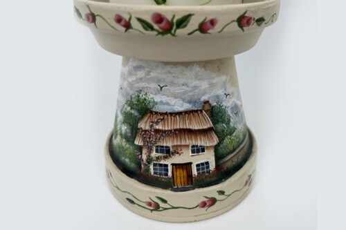 Ceramic vase with a hand-painted countryside scene: thatched cottage, trees, birds, under a cloudy sky, with a pink floral border at the top edge.