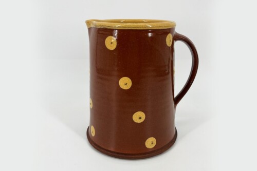 Brown ceramic pitcher with yellow polka dots on a light studio background, front view and handle visible