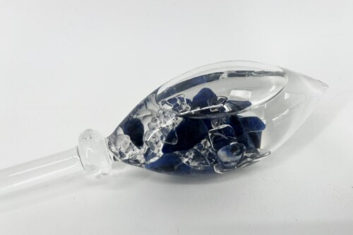 Clear glass bottle tipped on its side, filled with blue glass shards inside on a white background.