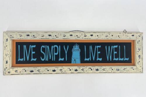 Framed decorative sign: navy panel reading 'LIVE SIMPLY LIVE WELL' in blue with a central lighthouse illustration, set in a white embossed frame.