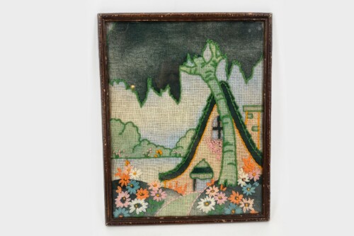 Framed needlepoint artwork showing a thatched-roof house beside flowering garden and green hills/mountains in the background.