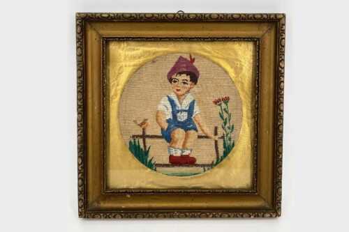 Embroidered round portrait of a young boy in blue overalls and a purple hat sitting on a bench, with flowers to the right, framed in a gold ornate frame.