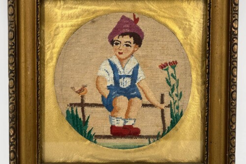 Cross-stitch embroidery of a young boy in blue overalls sitting on a fence with flowers nearby, wearing a purple hat.