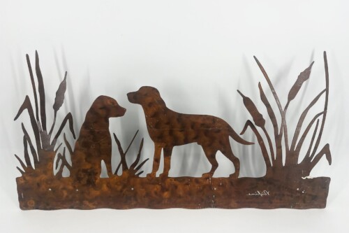 Rust-colored metal silhouette sculpture showing two dogs facing each other among tall cattails in a marshy scene, with a uneven base at the bottom and a signature on the lower right edge.