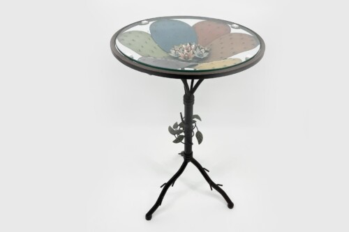 Round glass-topped side table with a pastel cactus-drawn surface on a black wrought-iron stand.