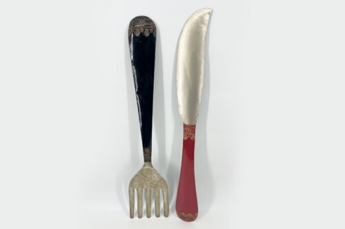 Three antique-looking utensils arranged vertically: a dark-handled knife, a curved silver-bladed knife, and a beige fork with a red-hued handle.