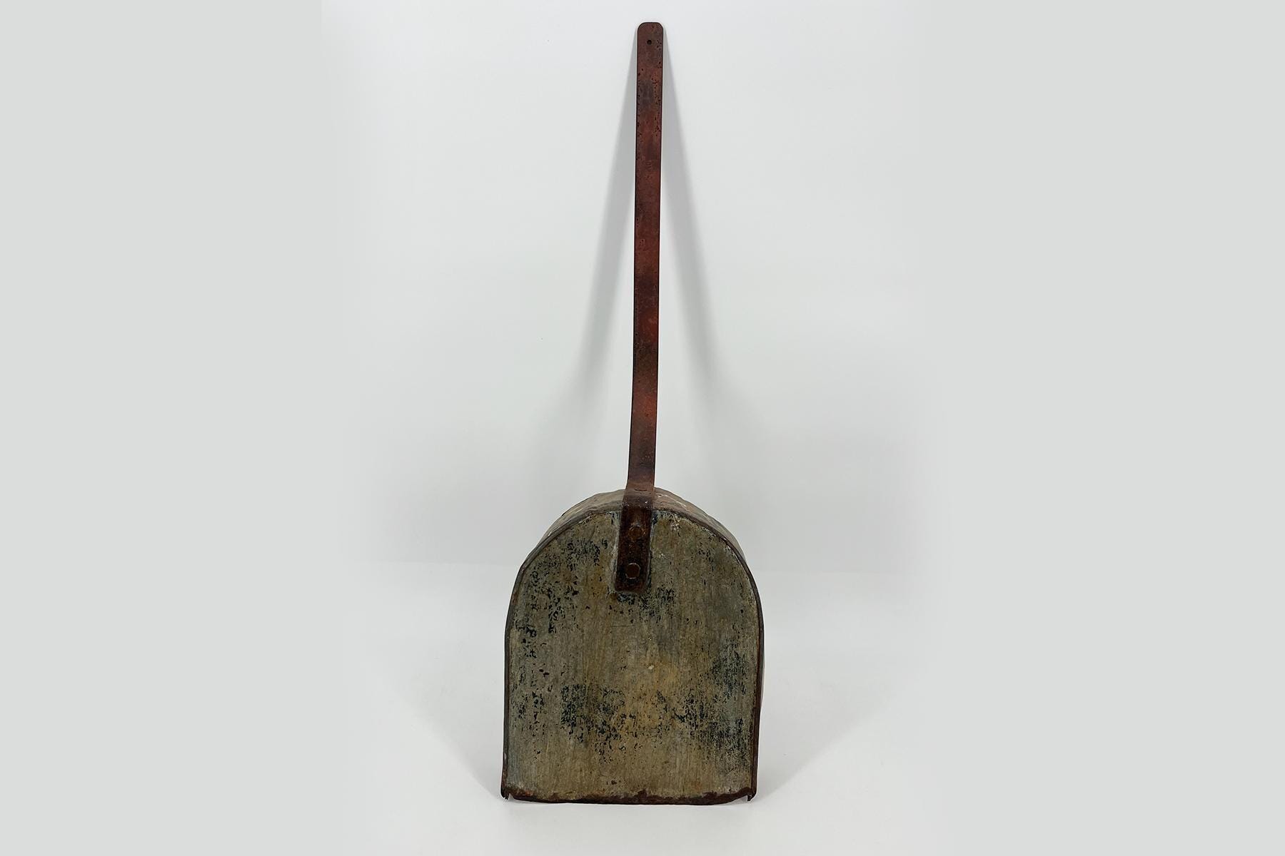 Antique, rusted metal tool featuring a long, narrow vertical handle attached to a rounded, weathered container on a plain background.