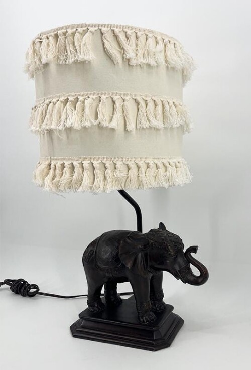 Table lamp with a black carved elephant base and a fringed cream lampshade.
