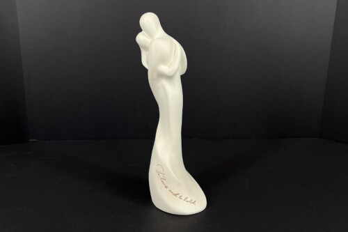 White abstract sculpture of a stylized standing figure with an upraised arm, mounted on a curved base with cursive lettering along its side (inscribed text partially visible).