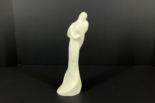 White abstract statue of a tall, robed human figure standing on a dark surface against a black backdrop.