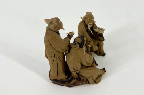 Three brown-clay figurines of elderly men in robes gathered around a central object, on a dark wooden base, against a white background.