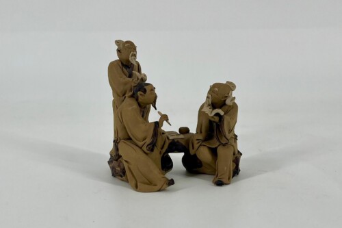 Three tan clay figurines in robes gathered around a low table, engaging in a scene of study or conversation on a white backdrop
