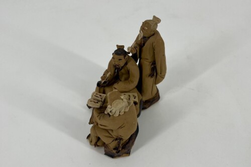 Three brown-clad clay figurines, two standing and one seated, gathered around a small, handheld object and looking focused.