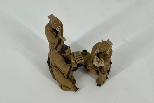 Three brown-clay figurines in robes are seated/kneeling and reading small books together, as if sharing a study or discussion.