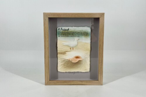Small paper artwork inside a wooden shadow box frame, showing a dove over a beach scene with the text 'A Friend'.