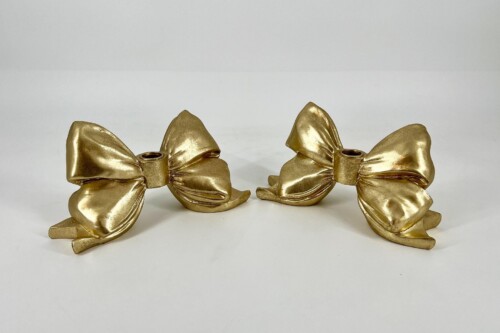 Two metallic gold bow ornaments lying on a white surface, facing each other in a minimal setup.