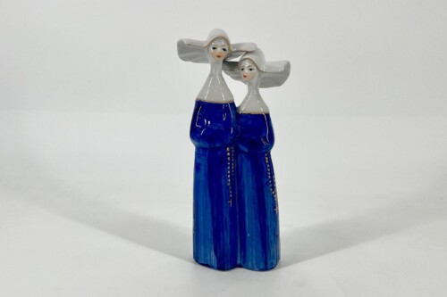 Ceramic sculpture of two women standing side by side, wearing blue dresses with white tops, facing forward with heads tilted.