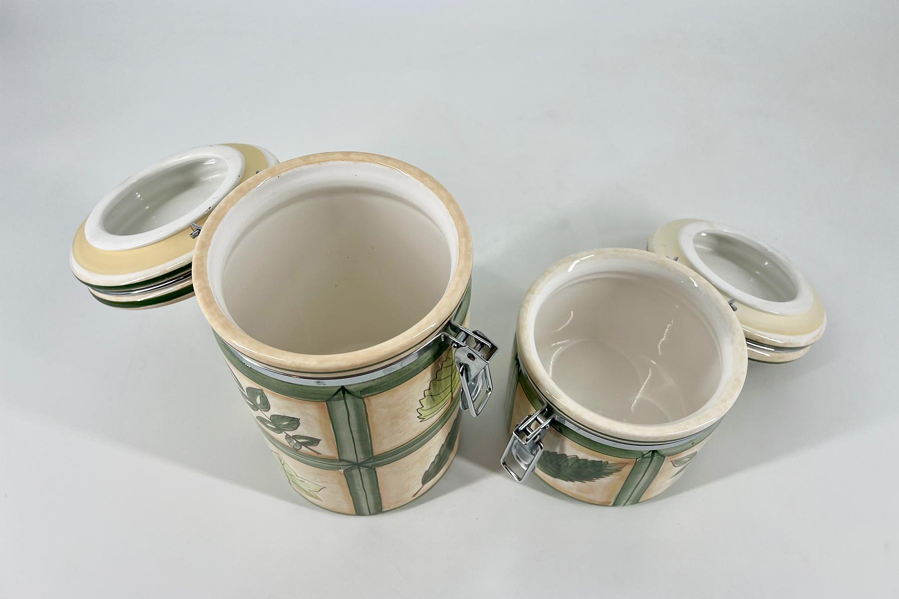 Three decorative ceramic jars with lids and metal clasps, featuring green botanical patterns on beige glaze, placed on a white surface.