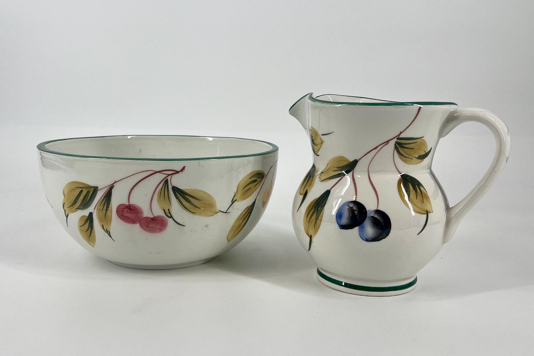 Two white ceramic serving pieces with fruit and leaf motifs: a wide bowl on the left and a curved pitcher on the right, both decorated with red cherries and green-yellow leaves.