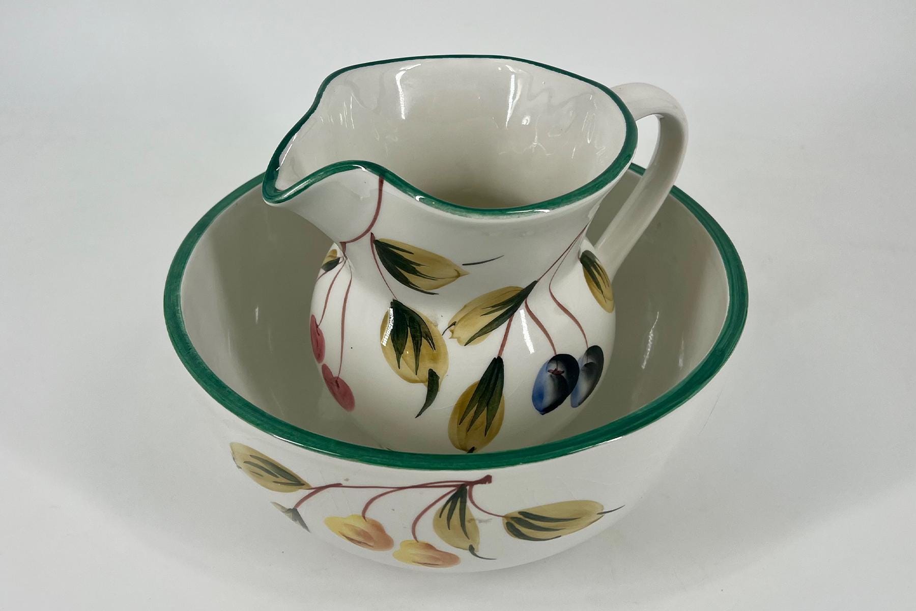 White ceramic pitcher with a green rim resting inside a matching bowl, both decorated with a yellow leaf and fruit pattern.