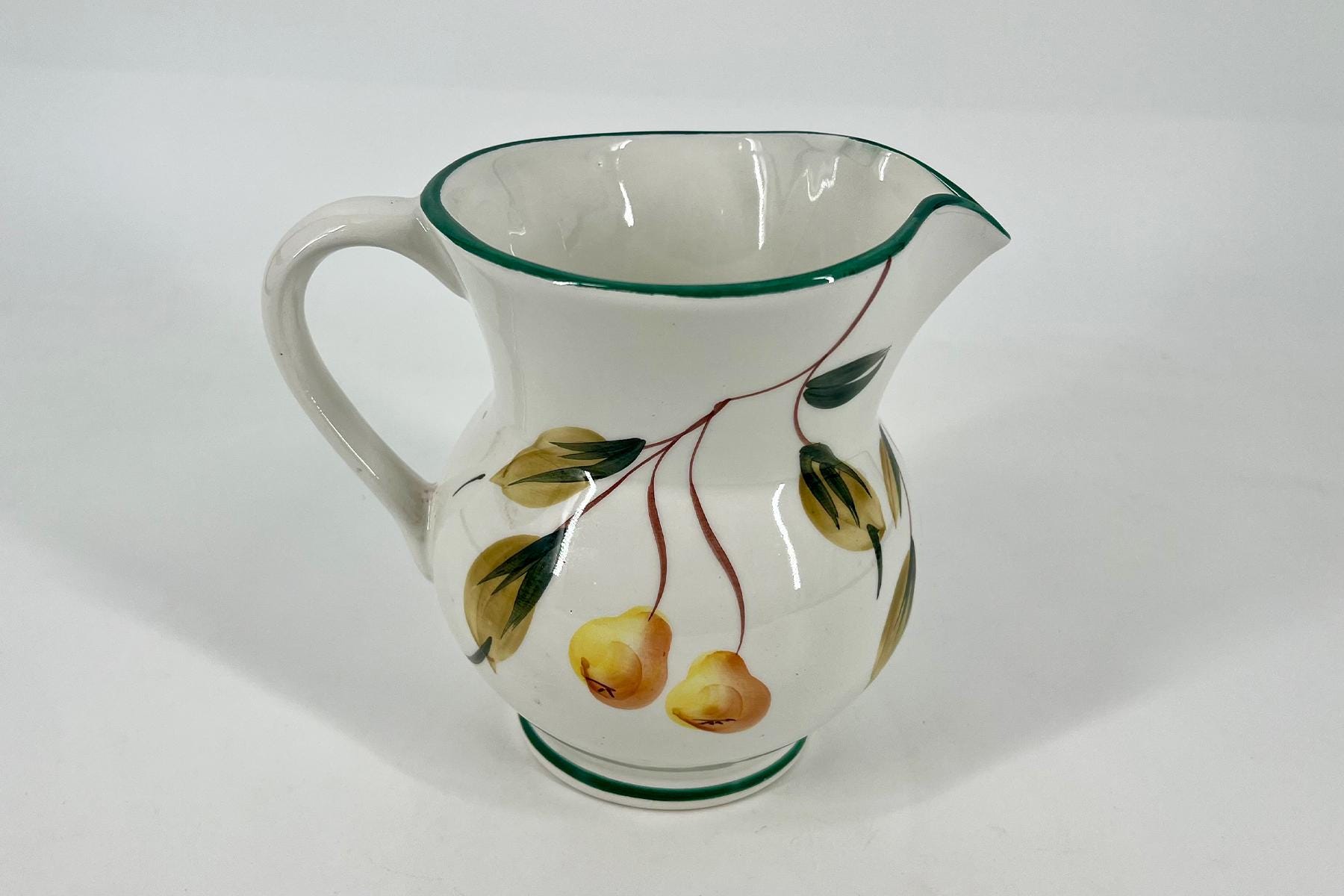 White ceramic pitcher with a green rim and handle, embellished with yellow fruit and vine decoration on a white background.