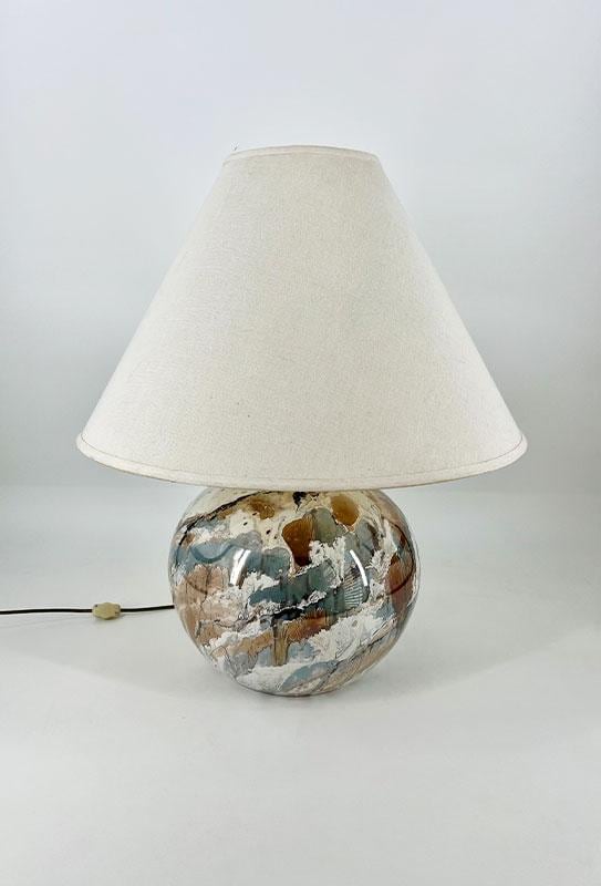 Table lamp with a round, marbled ceramic base in blue, brown, and white, topped by a beige fabric shade; cord extends to the left.