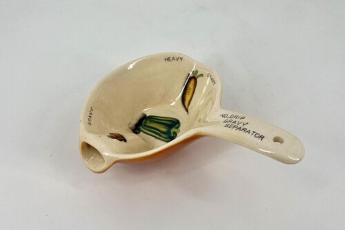 Ceramic gravy separator molded like a pelvic bone, with printed labels 'No-Drip Gravy Separator' and a carrot illustration inside.