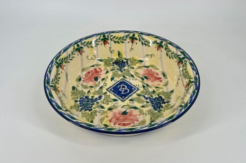 Top-down view of a decorative ceramic bowl with a blue rim, floral vines, pink roses, and grape clusters on a yellow background