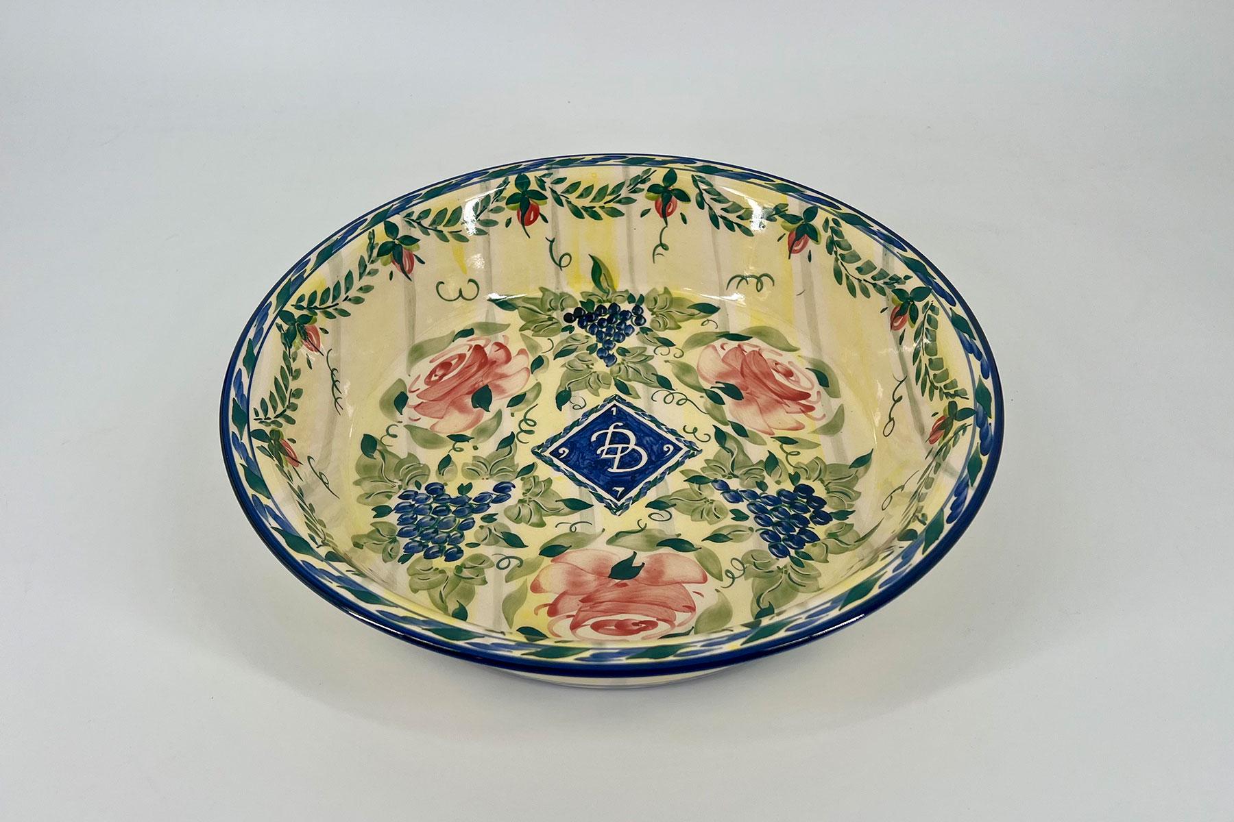 Top-down view of a decorative ceramic bowl with a blue rim, floral vines, pink roses, and grape clusters on a yellow background