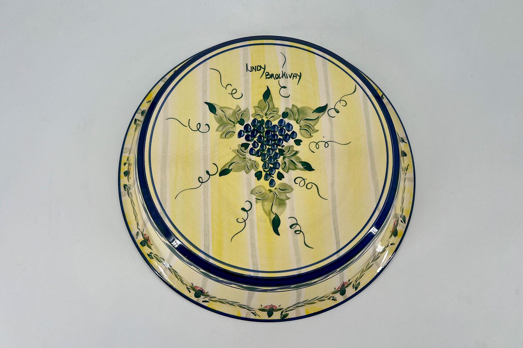Decorative ceramic plate with a yellow striped pattern, central grape motif, and a blue rim mounted on a light wall.