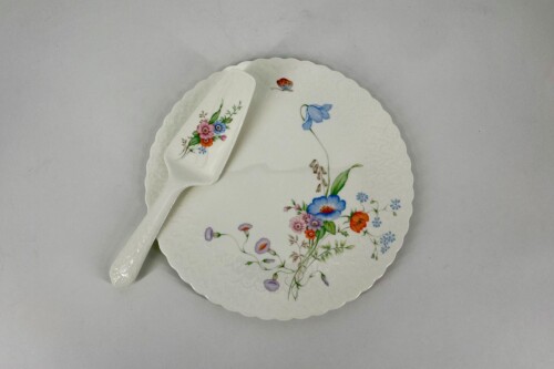 White porcelain plate with colorful floral pattern and scalloped edge, accompanied by a matching floral spoon on a gray surface.