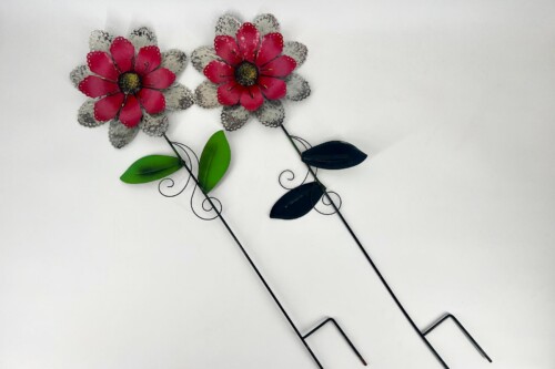 Two metal decorative flowers with pink-red petals, gray outer petals, green leaves, and curly wire stems against a light background.