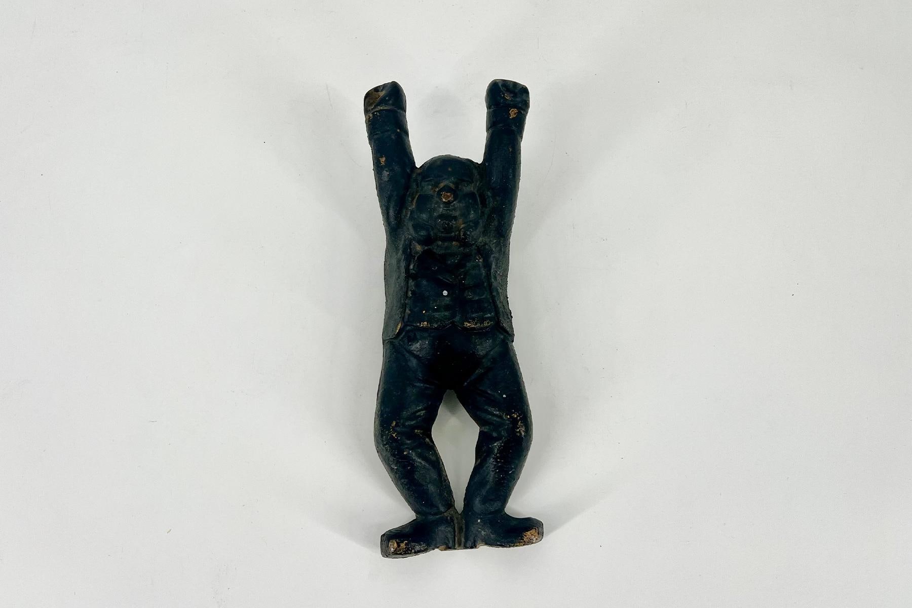 Small dark figurine of a human with arms raised overhead, standing on a flat base on a light background.