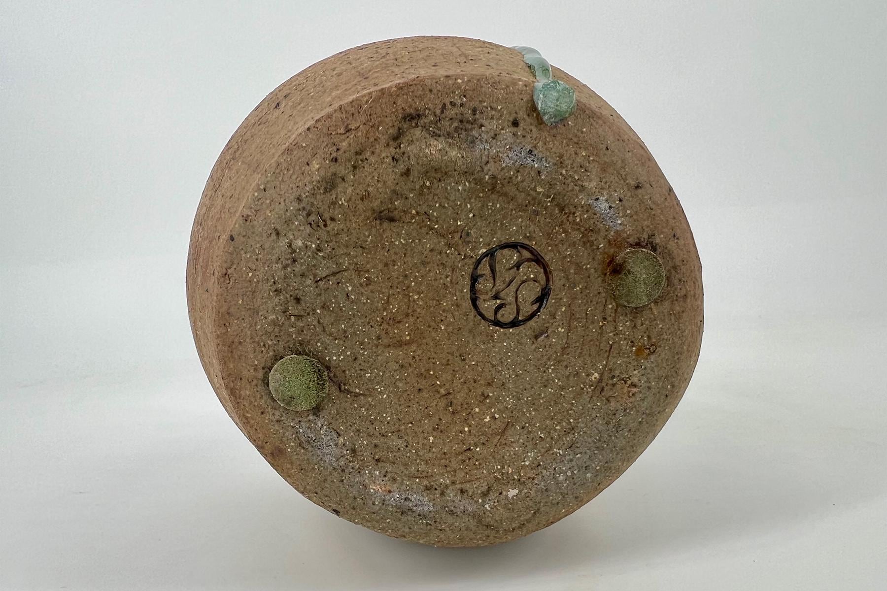 Close-up of the bottom of a textured, sandy-brown ceramic pot with small moss patches and a circular decorative emblem in the center.