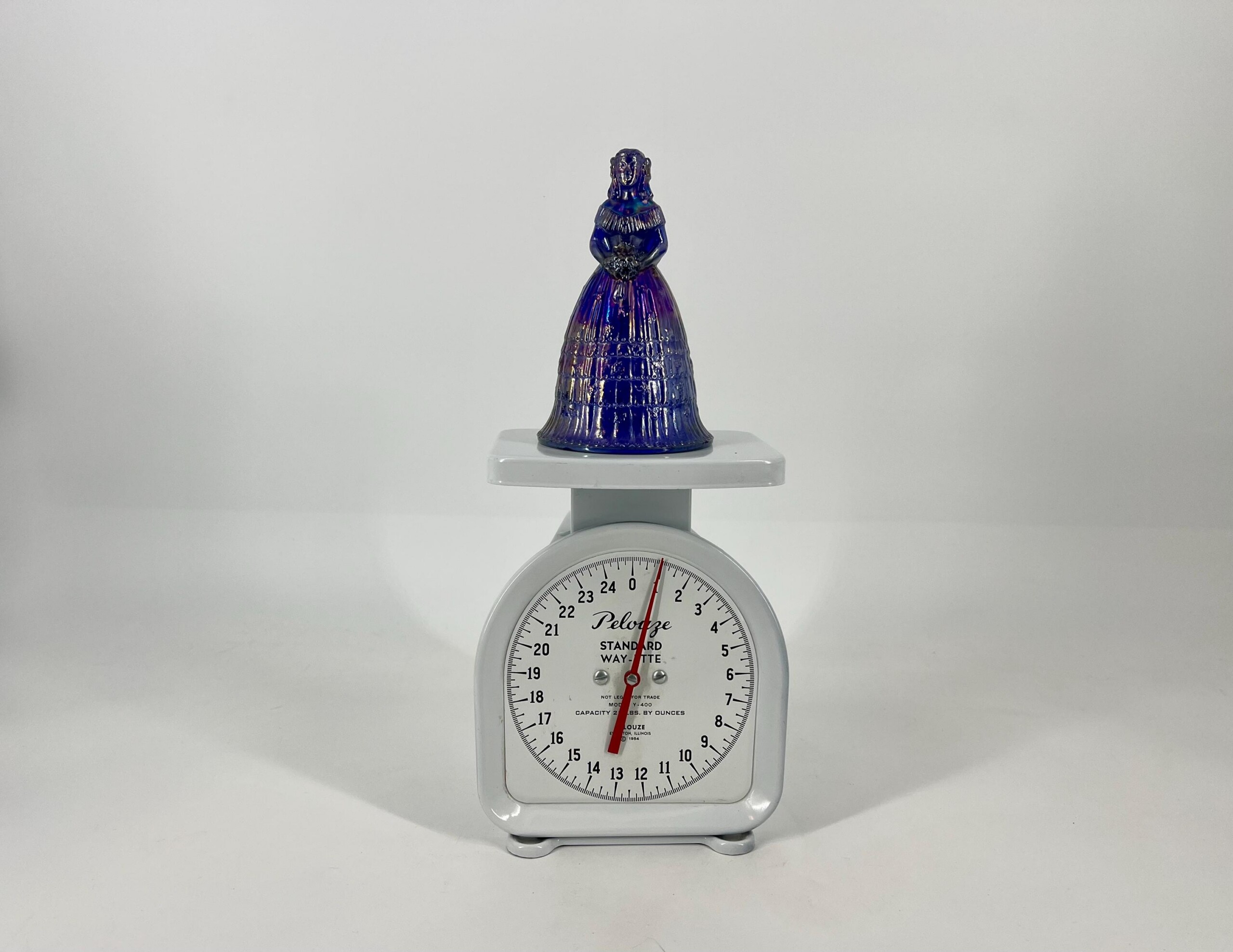 Cobalt-blue glass figurine of a woman in a long gown atop a vintage scale, against a plain white background.