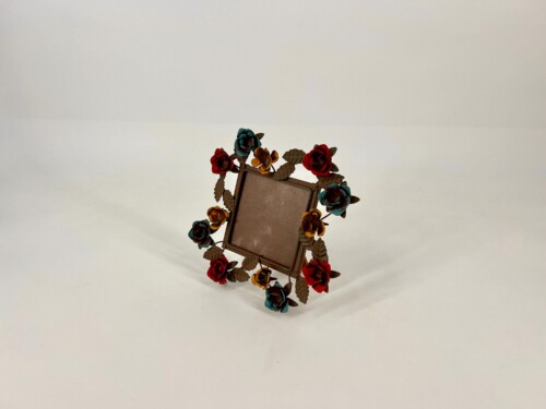 Tilted brown photo frame wrapped in colorful metal flowers (red, teal, yellow) against a pale backdrop, decorative sculpture.