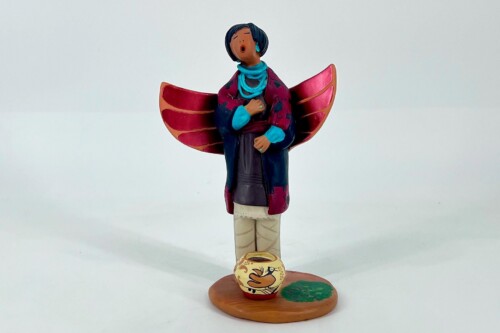 Ceramic figurine of a person with blue scarf and magenta jacket, large pink wings, standing on a wooden base with a painted vase nearby.