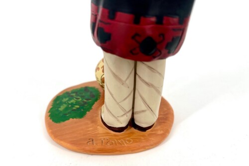 Back view of a small figurine standing on a round wooden base; beige pants with brown shoes and a red/black upper garment; green patch on the base.