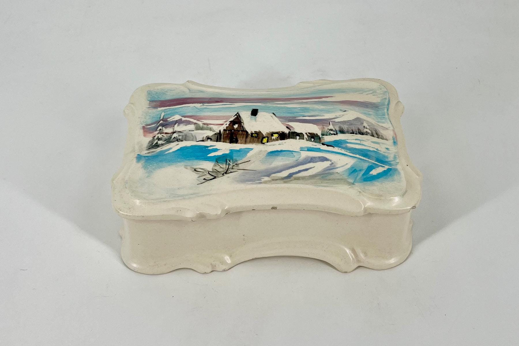 Ceramic decorative box with a hand-painted winter scene: snow-covered cottage, mountains, and blue sky on the lid.