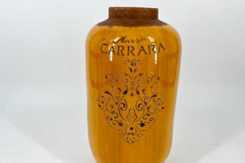 Orange-yellow ceramic vase with a dark brown rim and intricate scrollwork pattern; faint 'CARRARA' script near the top.