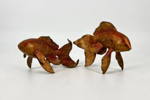 Two carved wooden goldfish sculptures in amber tones, with flowing fins, arranged on a white surface facing opposite directions.