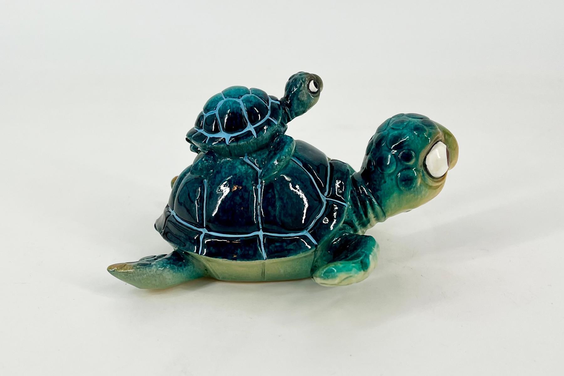 Two glossy ceramic turtles in teal and green tones; a larger turtle on the bottom with a smaller turtle perched on its back against a plain white background.
